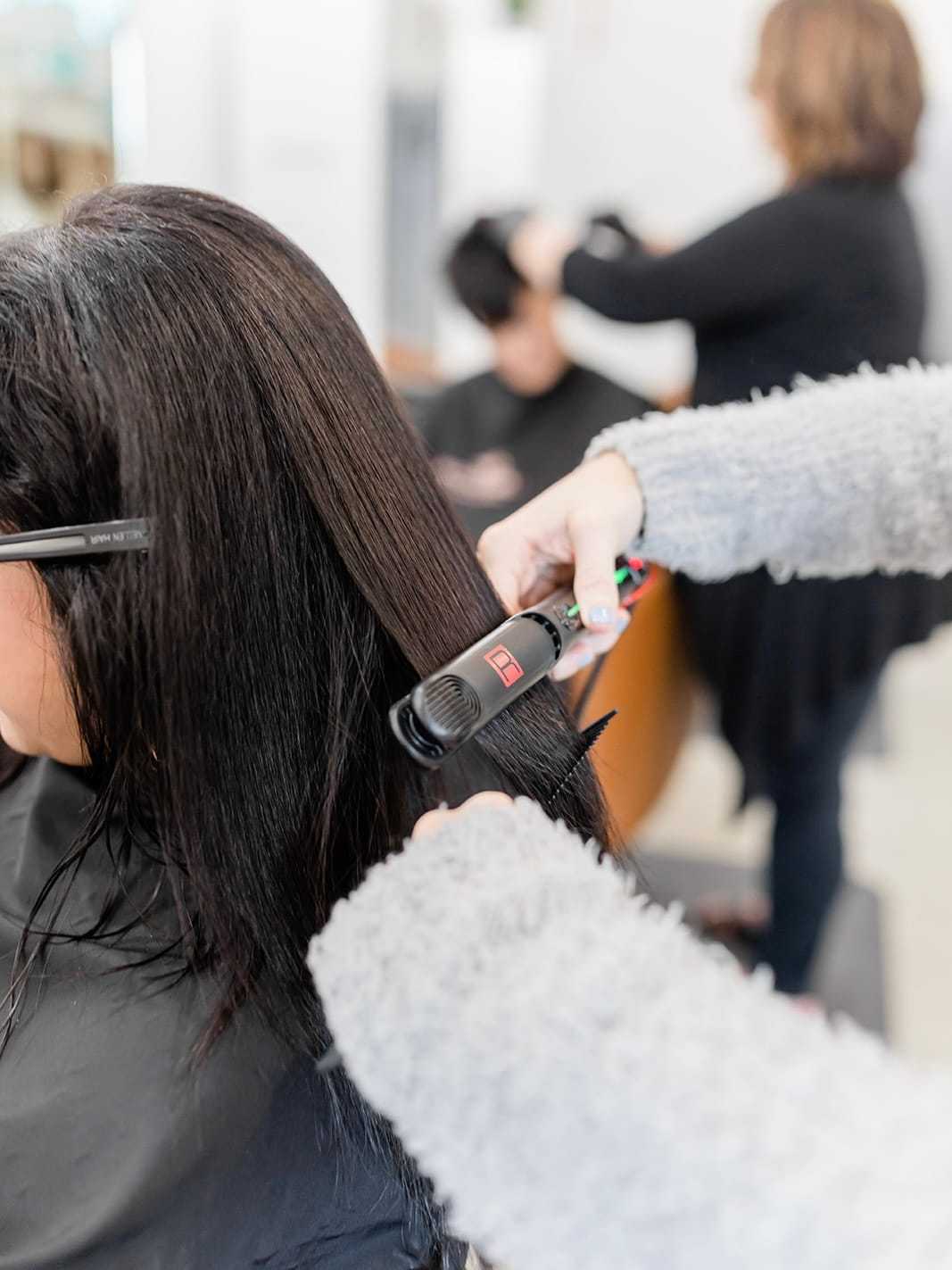 Hairdresser straightening a client's long dark hair in a salon setting.